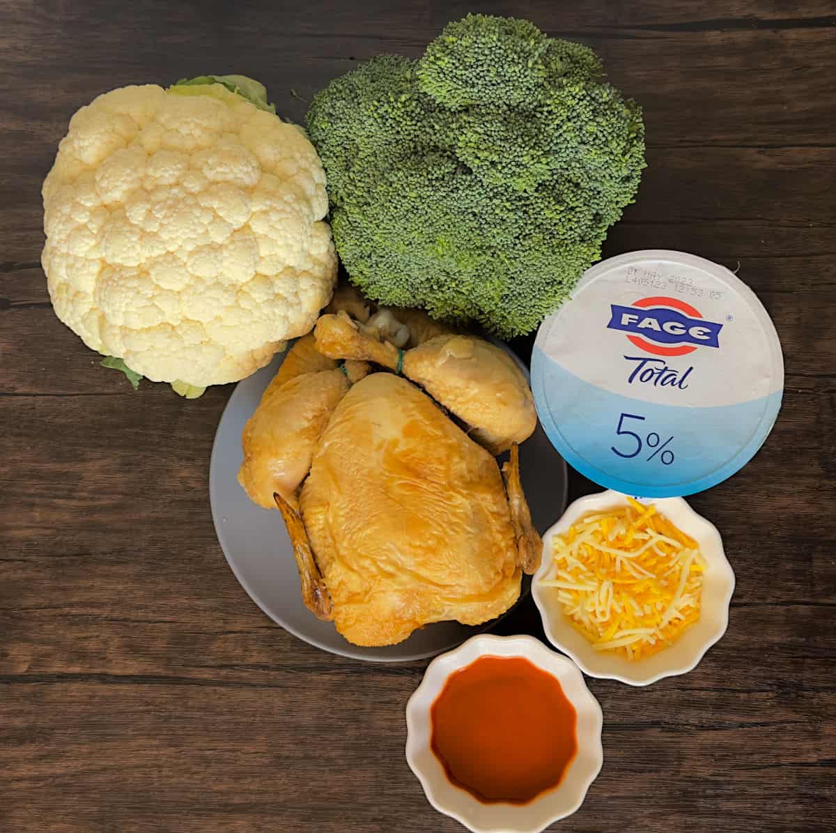 a wooden table with a grey plater and a whole rotisserie chicken on it surrounded with a white ramekin with red buffalo sauce, one with shredded cheese, a Fage 5% yogurt container, a head of broccoli, and a head of cauliflower in the ingredients for keto buffalo chicken broccoli casserole in the instant pot photo