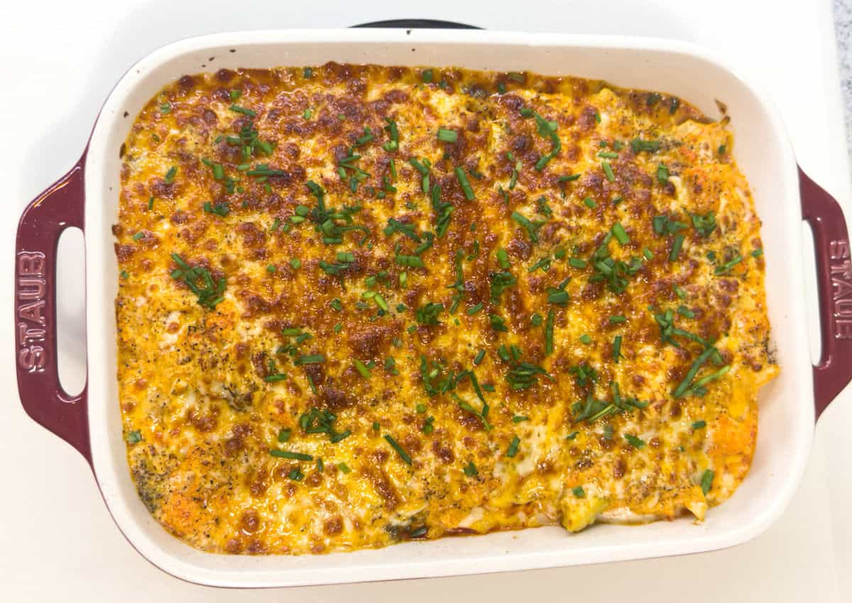 a red STAUB casserole dish with a cream interior holds finished product of just broiled keto buffalo chicken casserole with baked cheese on top slightly browned and garnished with chopped chives