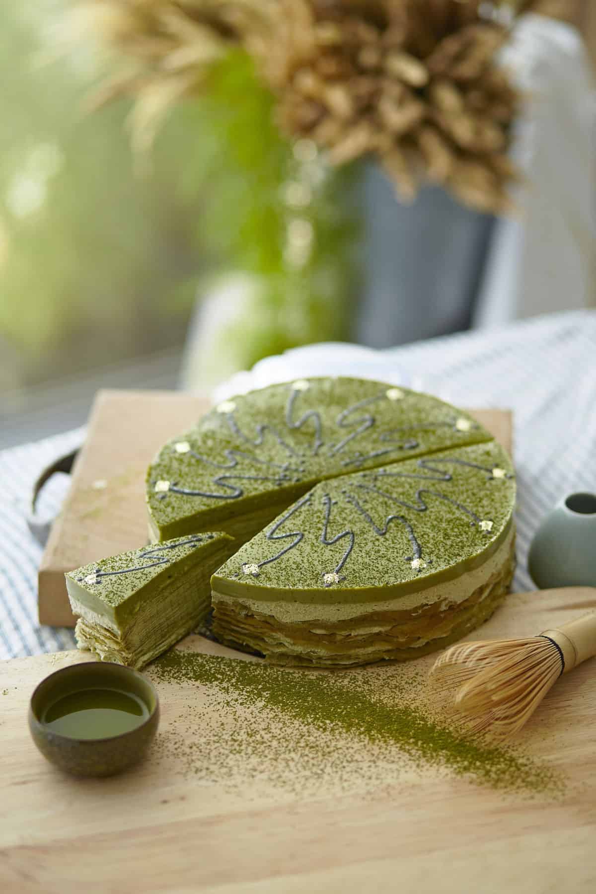 a light wood cutting board with green matcha powder around decoratively with a small dark colored cup of matcha on the left and a matcha whisk on the right in front of a beautifully baked layered cake with light green and white layers and decorative zig zag swirls on top of the cake to represent the culinary grade uses for matcha
