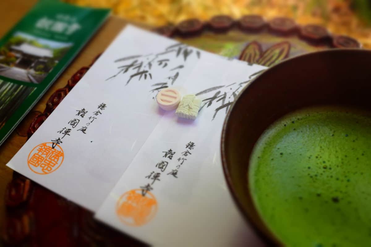 a close up photo of a traditional Japanese ceremonial matcha place setting with pieces of paper stamped with black and orange japanese writing and two candies next to a brown bowl in the right corner with vibrant green frothy matcha tea