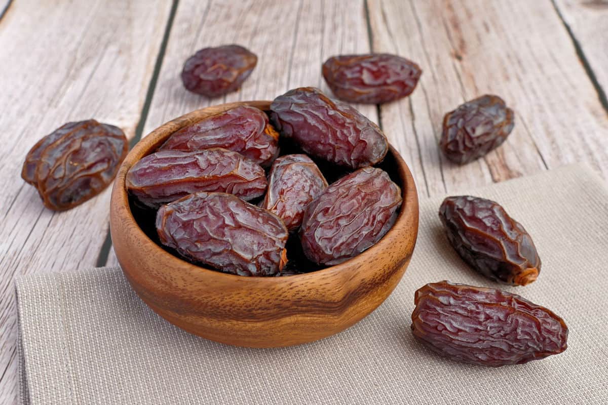 a tan wooden bowl with Auburn colored Medjool dates filled and falling around it decoratively on a lighter colored wood table and placemat of the same color