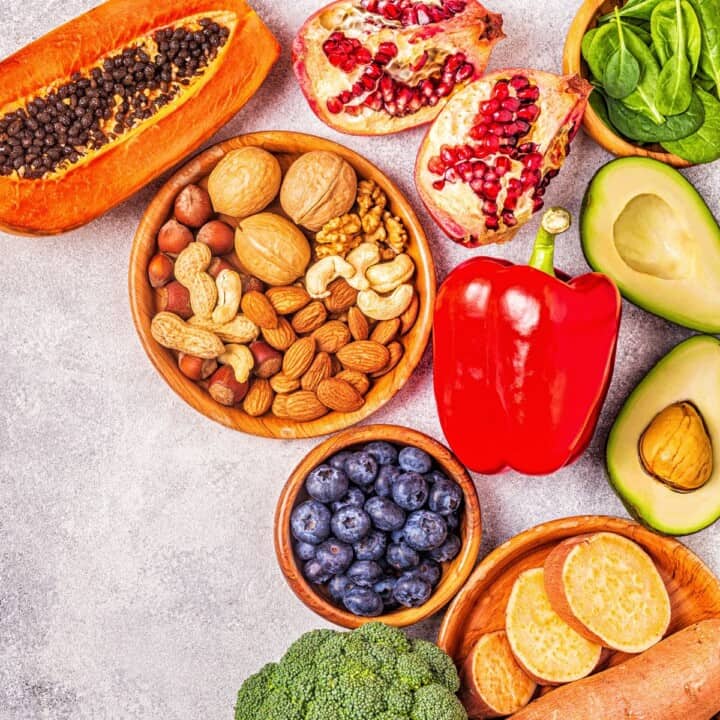 A closeup photo of anti-inflammatory foods like papaya, broccoli, root vegetables cut on a plate, cut avocadoes, blueberries, a pepper, pomegranate, and spinach in a bowl.