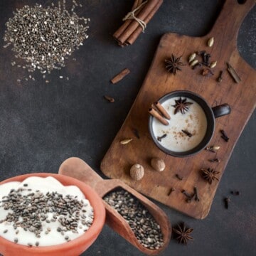 A dark background with a cutting board with several spices including chai and star anise nutmeg and cinnamon surrounding a dark mug of chai tea with a white liquid and spices in the cup including ground cinnamon and two cinnamon sticks. To the top left there is a pile of chia seeds and under that is an orange bowl of yogurt with chia seeds sprinkled on top and a wooden scoop full of chia seeds in it next to the bowl.