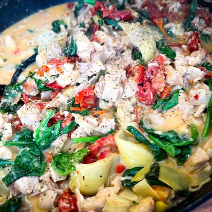 Creamy Tuscan Chicken cooking in the skillet as a close-up. Artichoke hearts, brightly colored wilted spinach leaves, sun-dried tomatoes, and chicken seasoned with pepper in a creamy sauce.