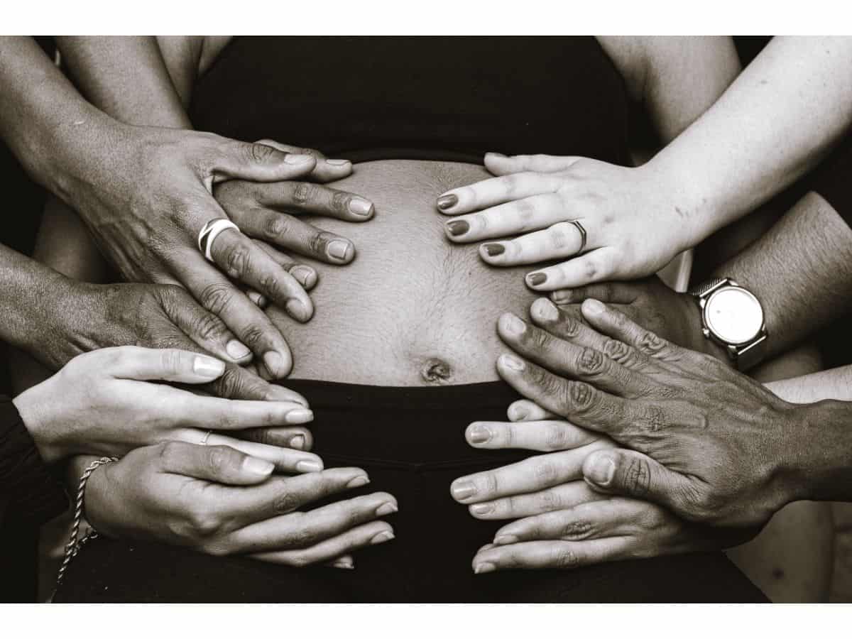 a sepia-colored photo of a woman with a crop top and several men's and women hands touching her belly