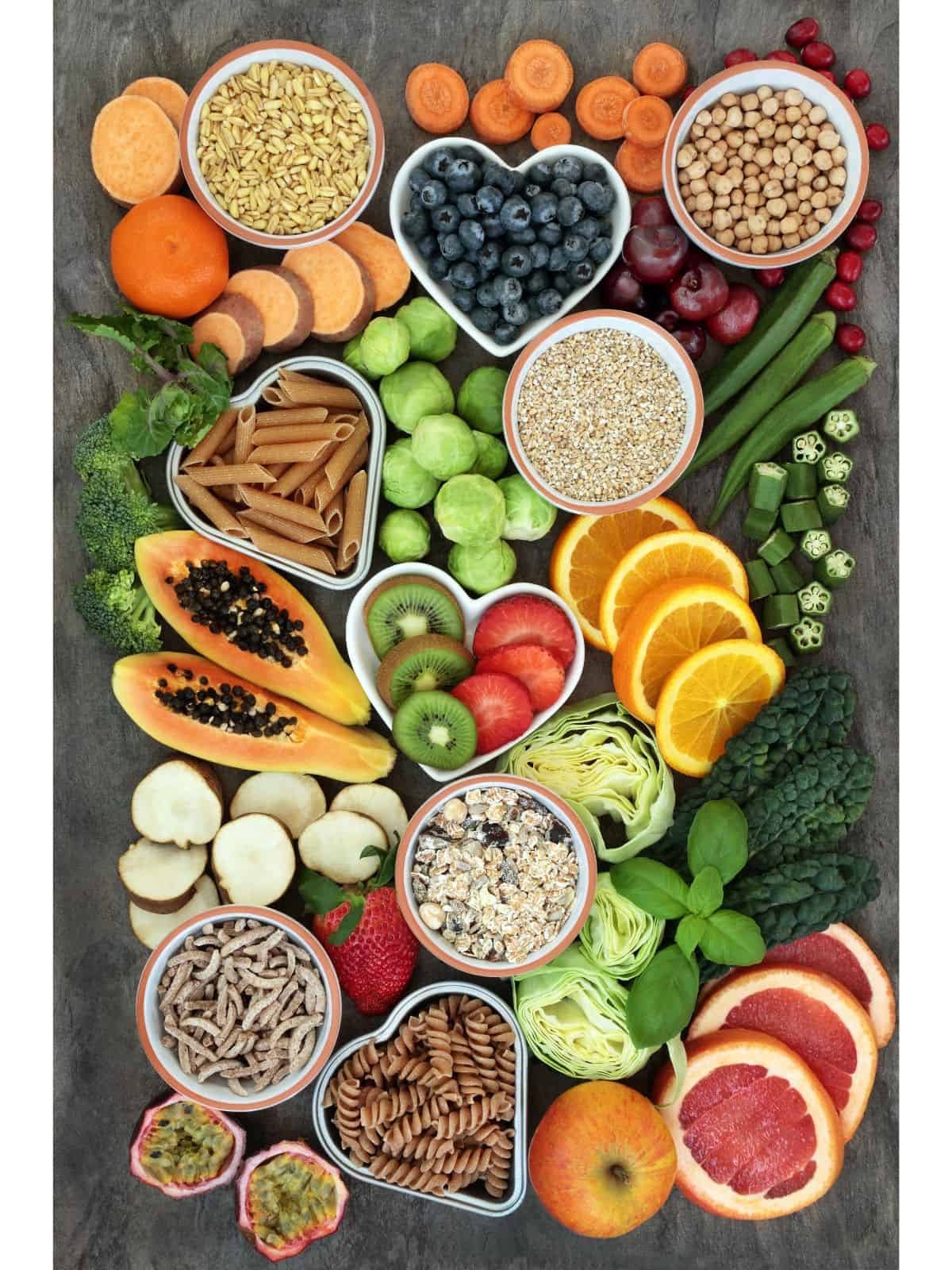 a decorative food design with several healthy and high vibrational foods in ramekins and cut up decoratively. These foods include pomegranate, apple, grapefruit, pasta, oats, cabbage, strawberries, kale, mint, moringa, okra, orange, papaya, kiwi, broccoli, brussel sprout, sweet potato, carrots, nuts, and seeds