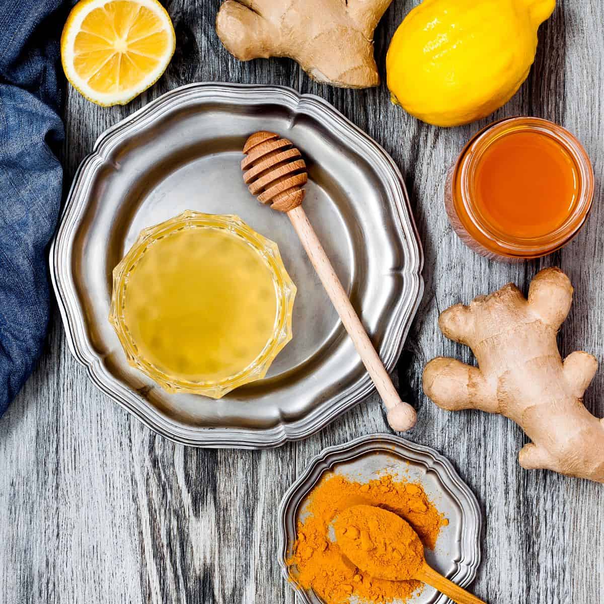 distressed wood with decorative silver platters with turmeric powder in a wooden spoon, lemon juice in a glass bowl, cut lemons, ginger, and a pot of honey and honeycomb stick. with a jean fabric in the side frame