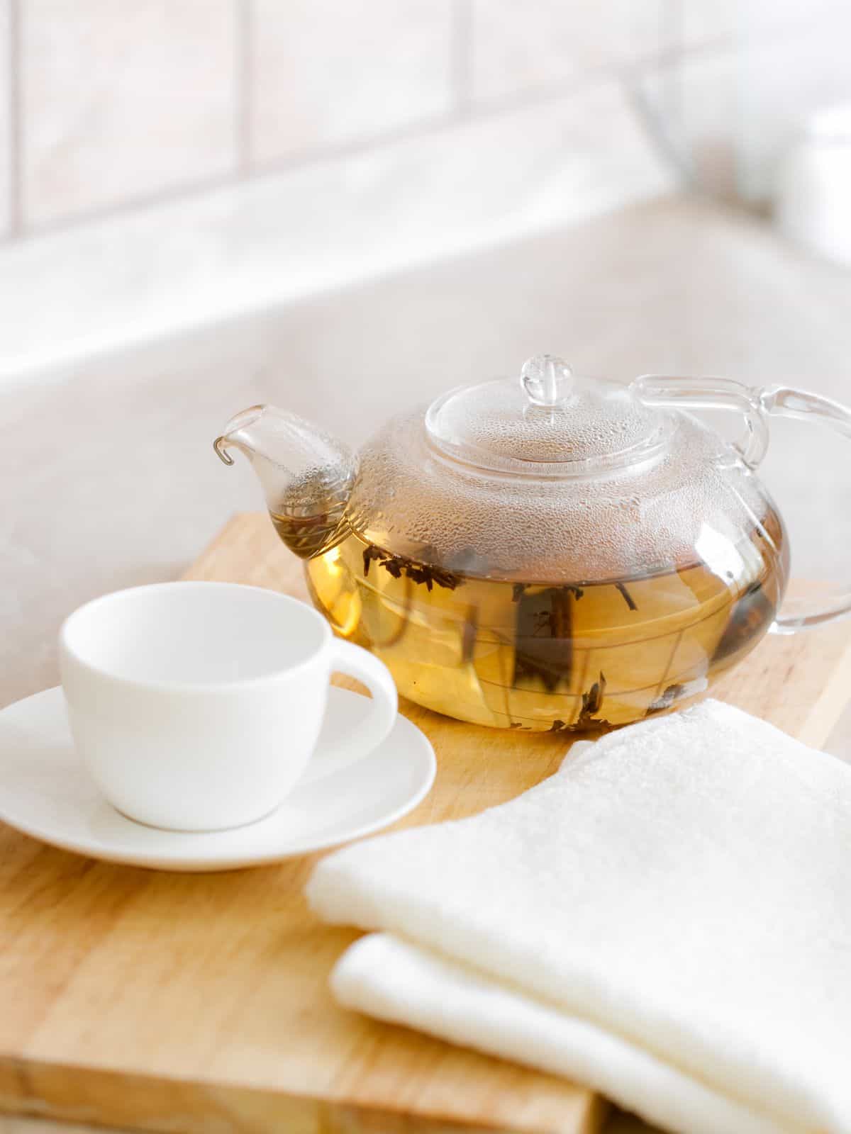 A clear glass tea kettle with light colored tea in it next to a white teacup on a light wood cutting board and napkin