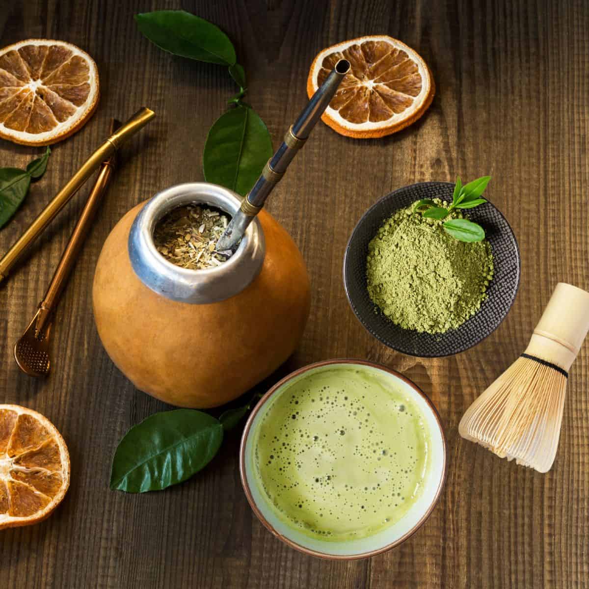 A wooden table with a burnt orange yerba mate gourd mug with silver lip and straw with herbs inside surrounded by dried orange slices and green leaves with special sifting tools laid next to it. on the right is a small brown-rimmed white cup of frothy matcha next to a bamboo whisk and black speckled bowl of vibrant green matcha powder