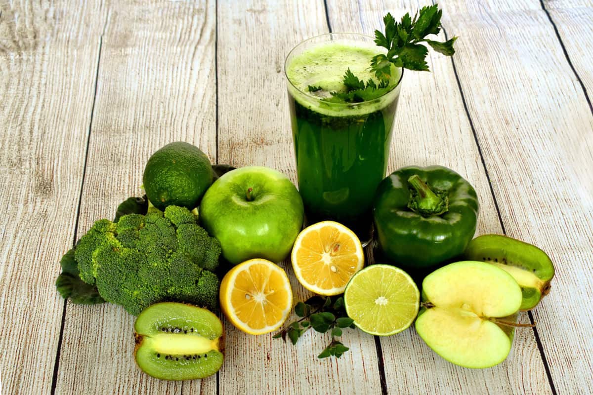 a wood-planked table going vertically with a presentation of a dark green juice in a clear glass with cilantro leaves to garnish on foam at the top of the glass. It is surrounded by broccoli, lime, green apple, green bell pepper, half of a green apple, two halves of lemon, two halves of kiwi and one half of a lime