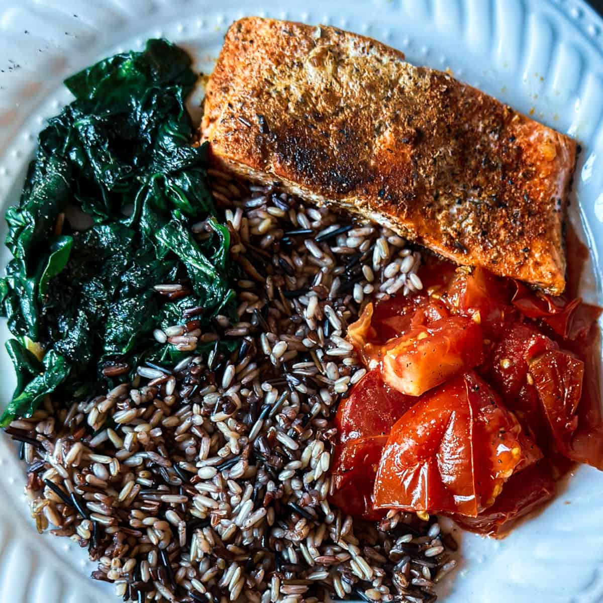 a photo of a white plate with a portion of lightly blackened salmon with cajun spices at the top of the plate with a bed of wild rice blend, sauteed spinach, and lightly cooked tomatoes cut into pieces.