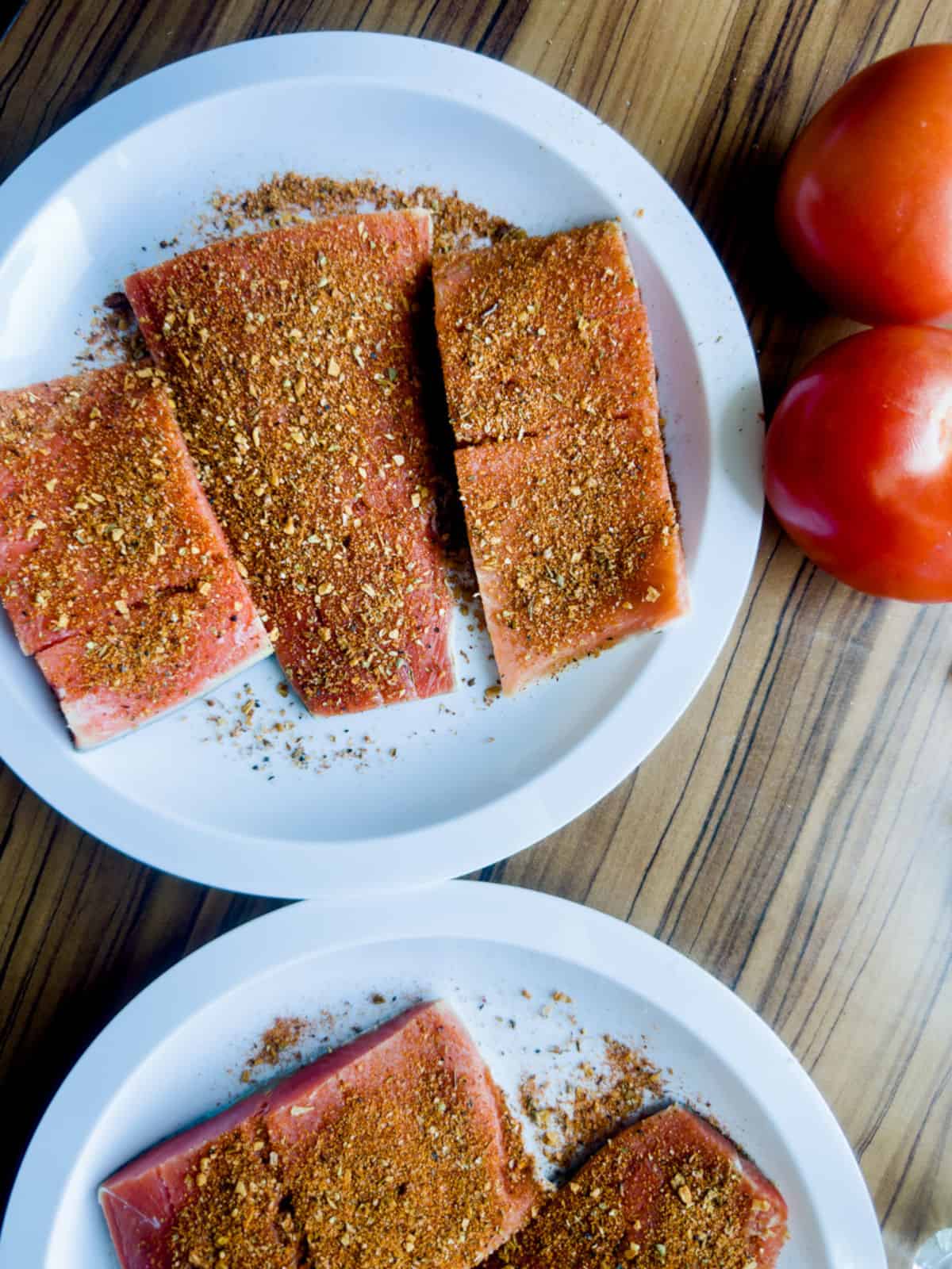 pink raw salmon portioned and seasoned and placed on two white plates with two tomatoes at the top right