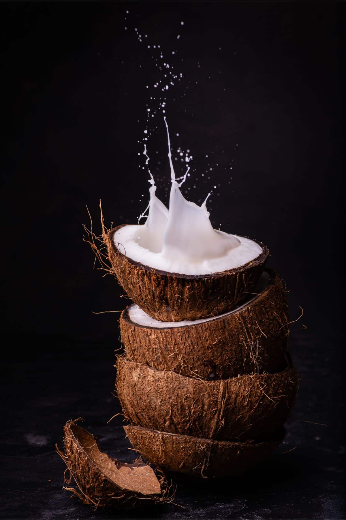 a decorative photo with several halves of coconuts cracked open stacked vertically on top of one another and a splash of coconut milk extending upward from the top half with a black background