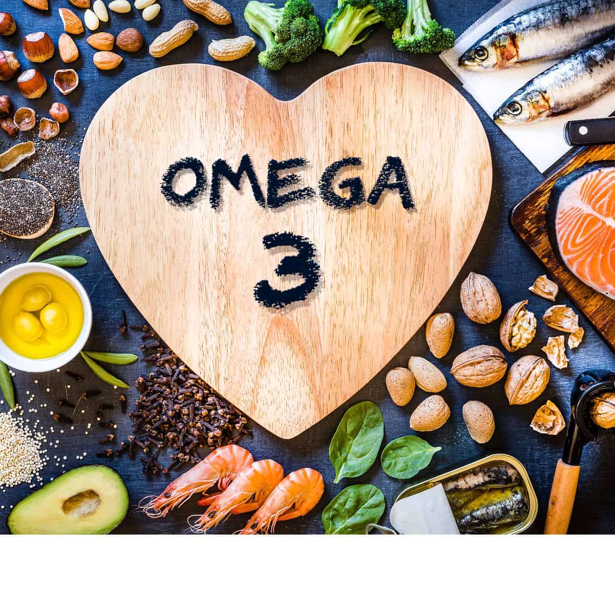 a wooden heart in the middle with black writing that reads omega 3. the heart is surrounded by seafood, spinach, sardines, avocado halves, olive oil, seeds, nuts, and salmon
