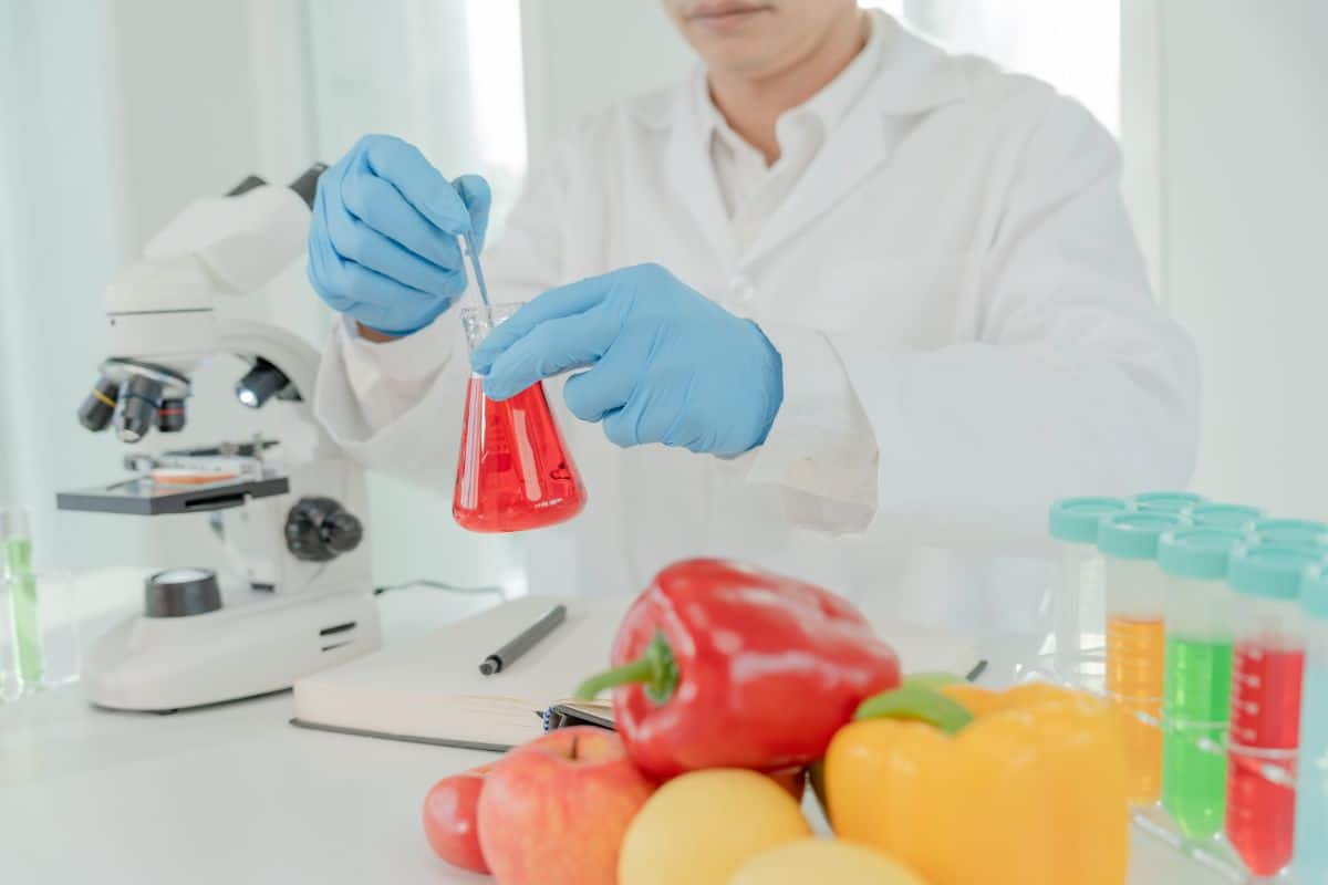 a photo of a scientist with a white coat and blue gloves with a beaker filled with red liquid. They are standing next to a microscope and various fruits and vegetables