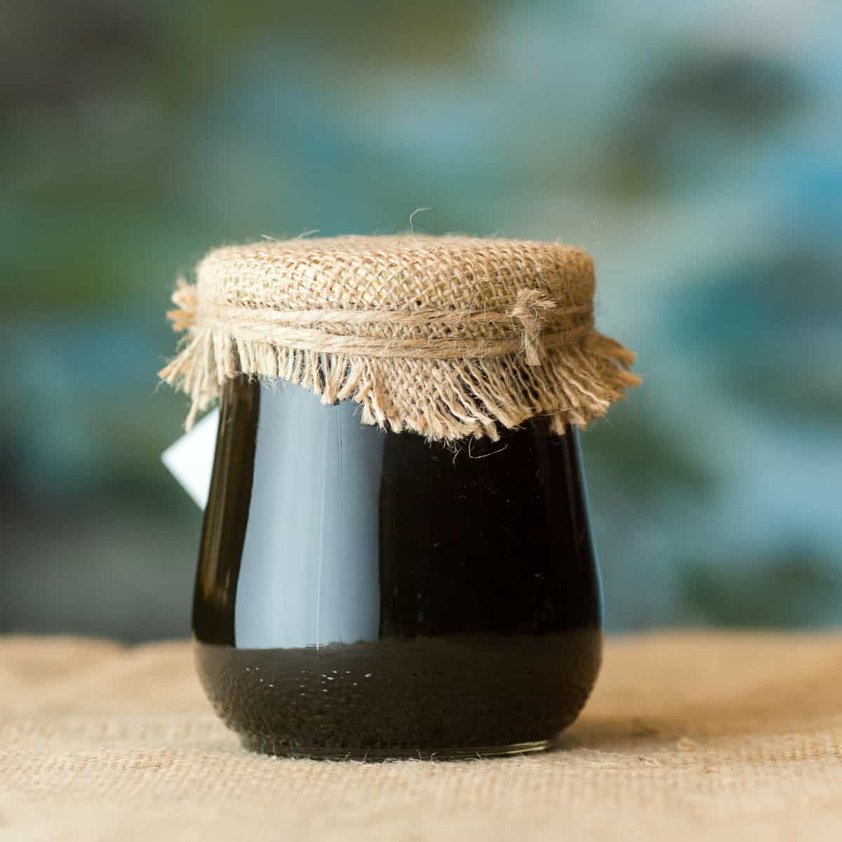 A simple photo of a glass jar of dark honey indicating buckwheat honey. It is covered with a burlap material.