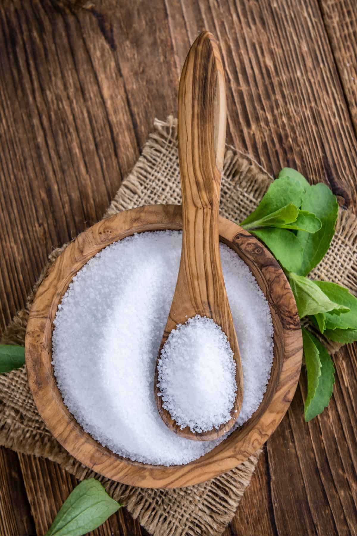 A wooden bowl and spoon with white stevia extract powder on top of a burlap cut out and surrounded by stevia leaves