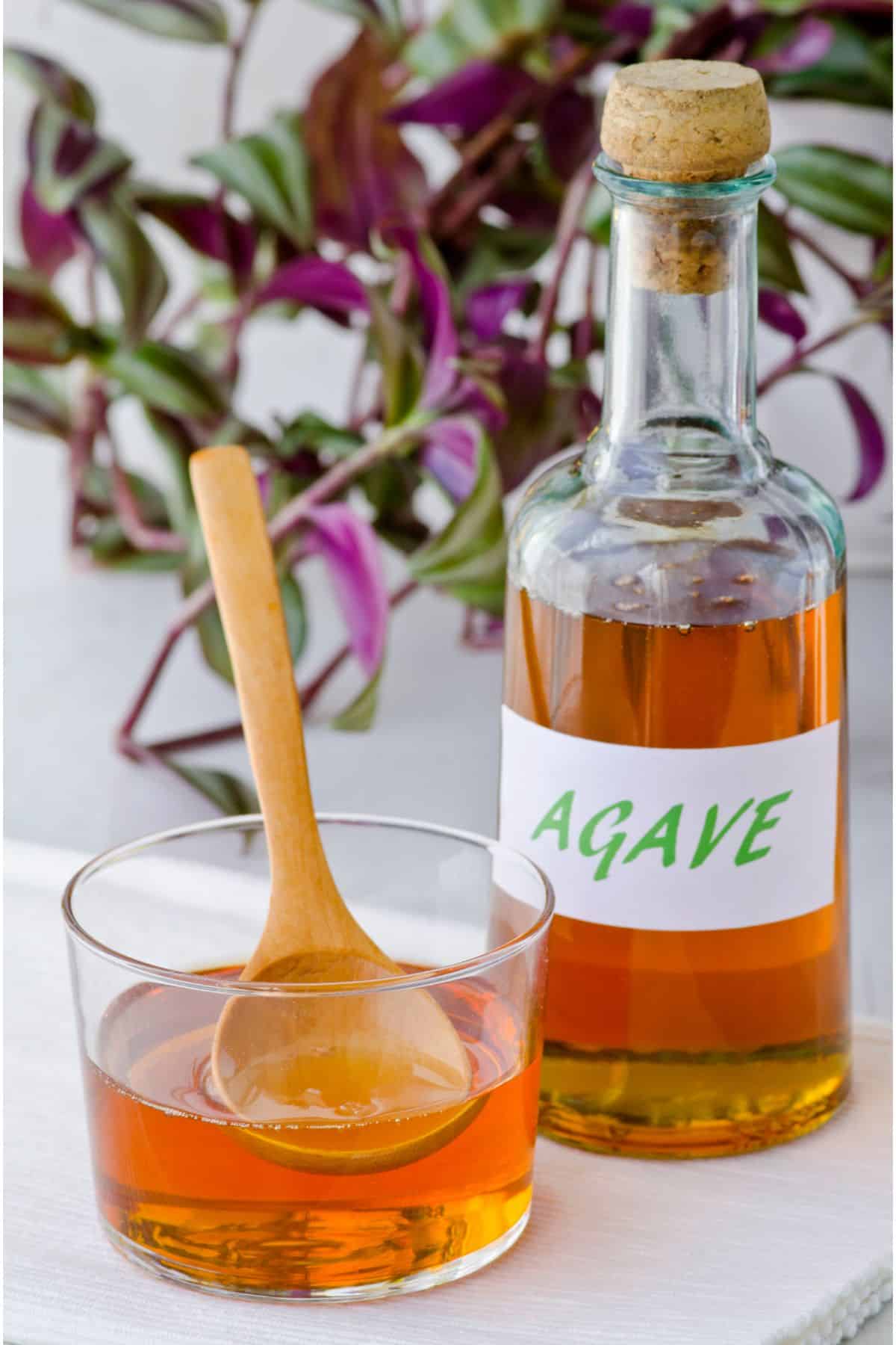 a light amber-colored syrup in a round rock glass with a light wooden spoon in it. Next to the glass is a taller bottle with the same amber liquid in it and a white label with green writing that reads AGAVE and a cork in it. In the background is green and purple flowers placed decoratively.
