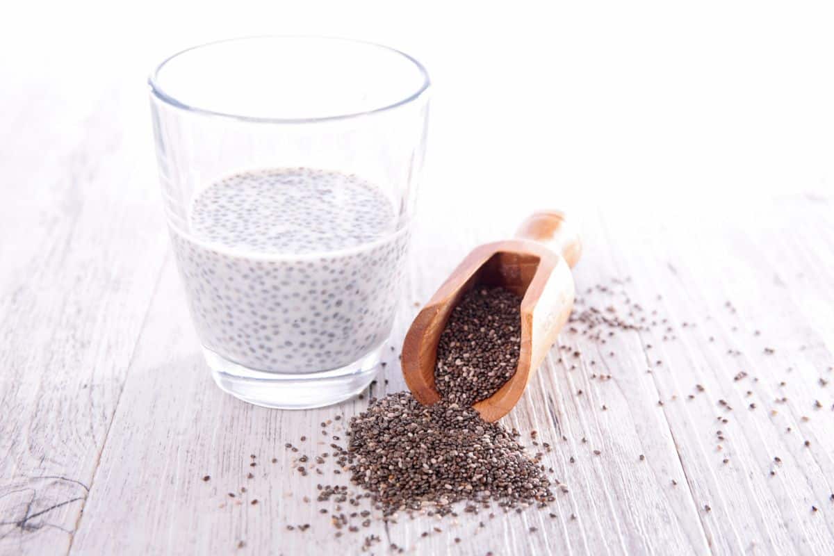A light grey wooden table with a dark wooden scoop with grey and white chia seeds spilling out of the scoop. Next to it to the left is a clear glass half full with milk and hydrated chia seeds.