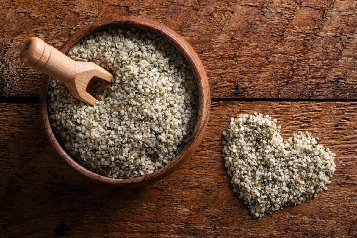 a brown wooden table with a wooden bowl filled with light colored hemp hearts and a pile of hemp hearts next to the bowl in the shape of a heart.