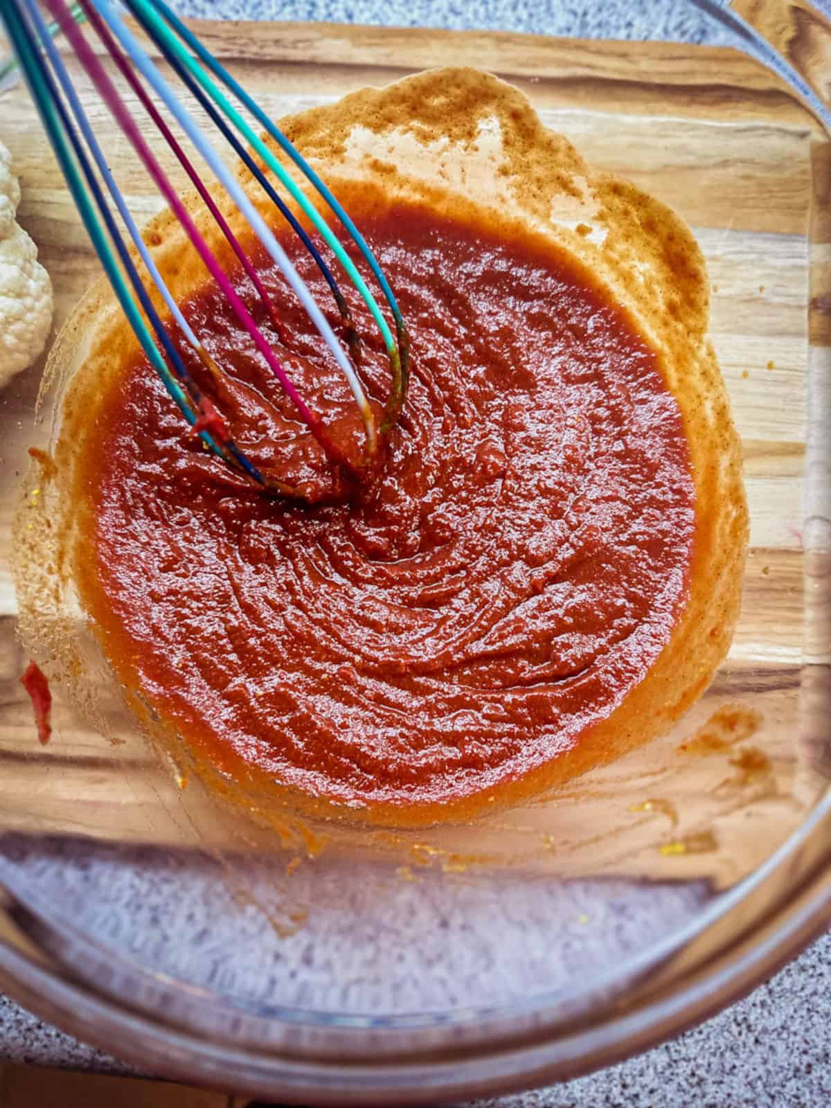 Close-up photo of barbecue sauce being mixed together in a glass mixing bowl with a multi-colored whisk. The bowl is set on a bamboo cutting board