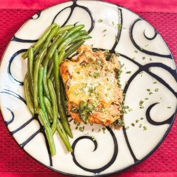 A red placemat with a white plate with black swirl decorations and outline with french green beans next to a square of keto BBQ chicken casserole garnished with chives and black pepper.