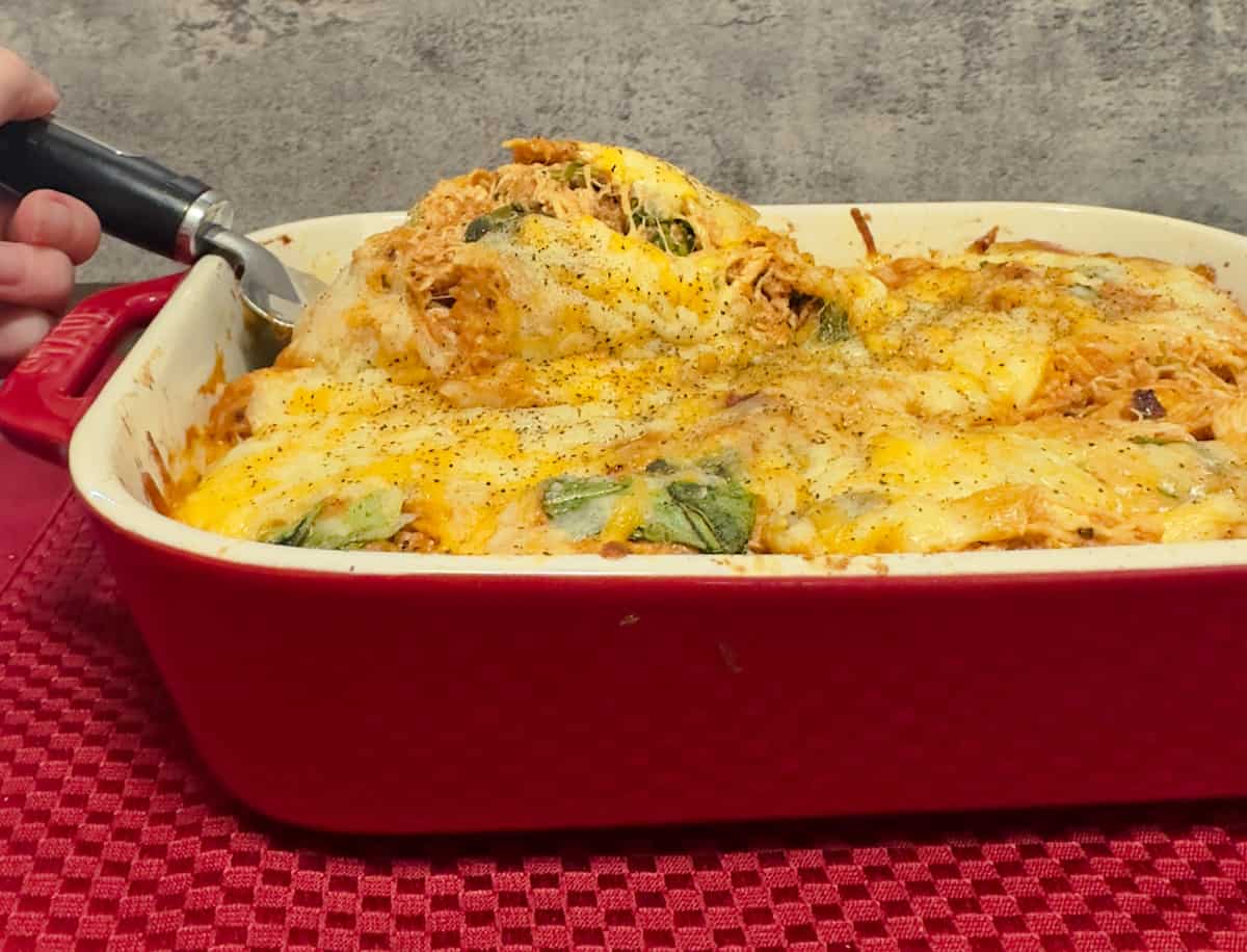 A red Staub casserole dish on a red placemat with baked keto BBQ chicken casserole garnished with black pepper. A serving utensil is beginning to scoop out a serving.