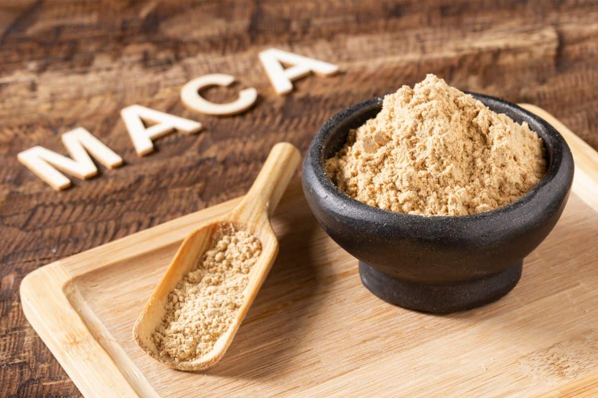 A decorative photo of a light wood bamboo cutting board on a dark wood table. The table at the top left has 4 wood cut out letters that say MACA. On the cutting board a bamboo scoop is placed with lightly colored powder maca in it. It sits next to a round black stone bowl filled with light fluffy powdered maca.