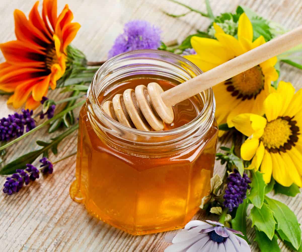 A decorative photo of honey jar with golden honey spilling out of it and a tan honey comb inside it. The honey jar is surrounded by various flowers of purple, white, yellow, and orange colors all on a wooden table.