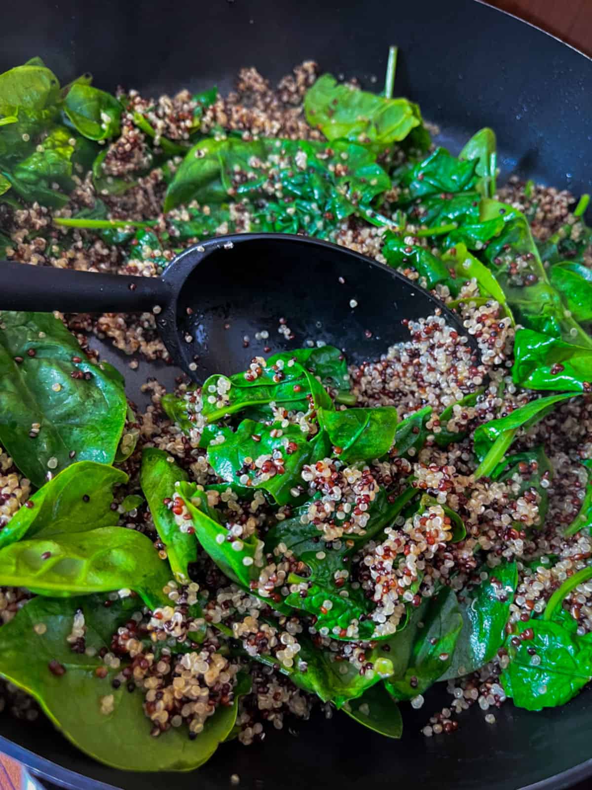 A black skillet and spoon with cooked tri-color quinoa with cooking fresh spinach being stirred in