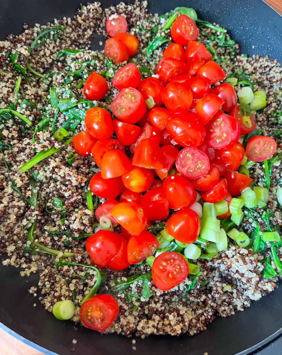 A black skillet with cooked tri color quinoa with cooked spinach, halved cherry tomatoes, and chopped green onions