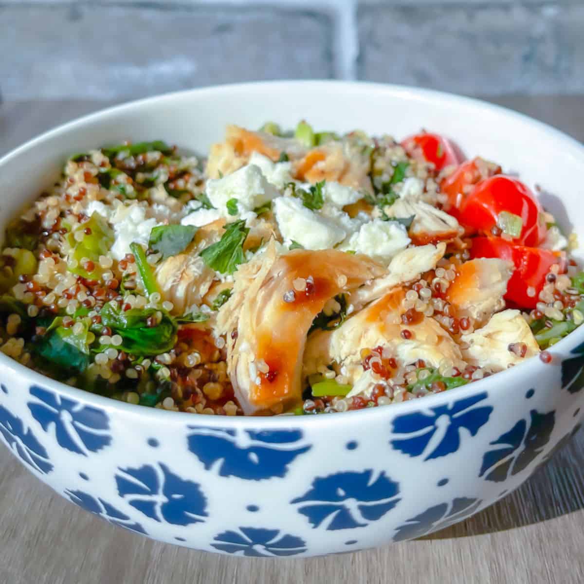 a white bowl with blue flowers painted on it with pesto chicken quinoa bowl mixed together quinoa and chicken mixture with feta tomatoes spinach, basil and basil pesto sauce