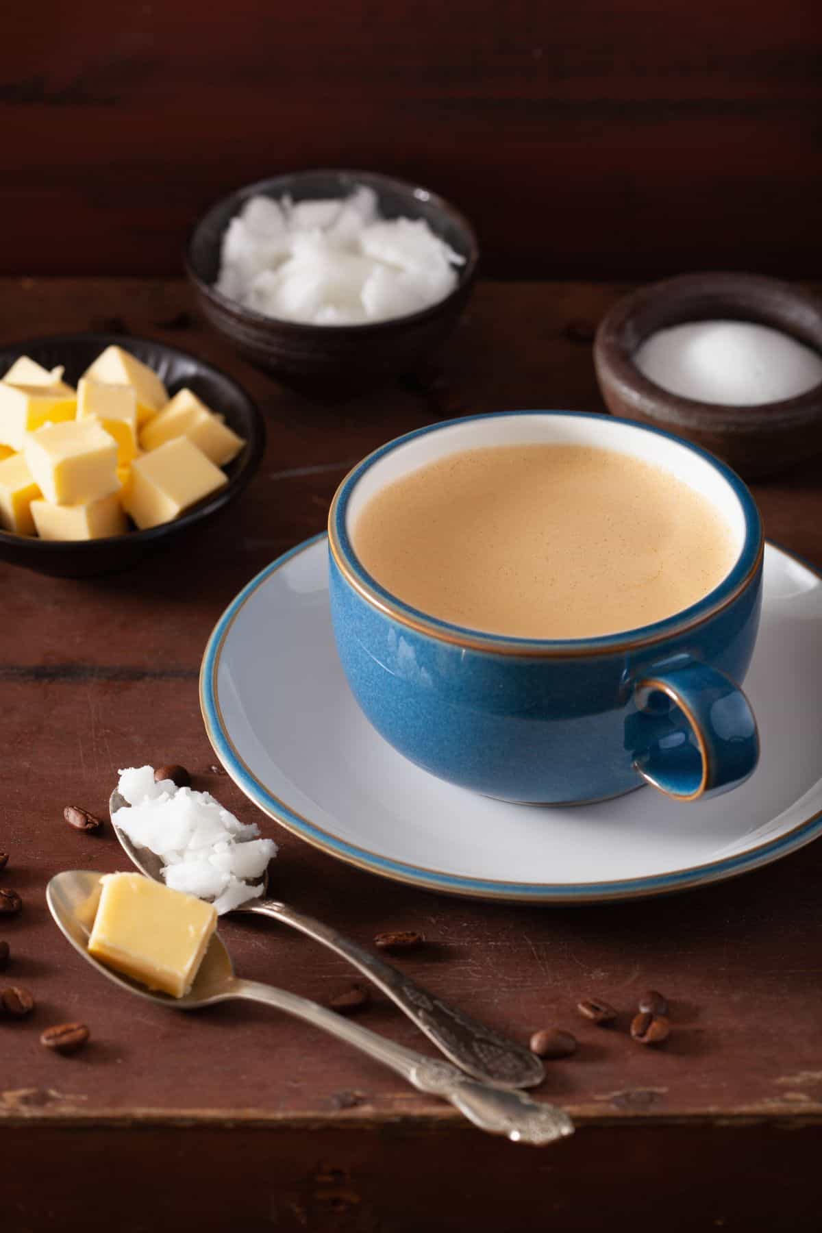 This photo illustrates the ingredients in bulletproof coffee. There is a dark wooden table with a blue cup of bulletproof coffee on a white saucer. Surrounding it is gradd-fed butter in a spoon, MCT coconut oil in a spoon, a bowl of ghee, a bowl of coconut oil, and a bowl of monk fruit sweetener.