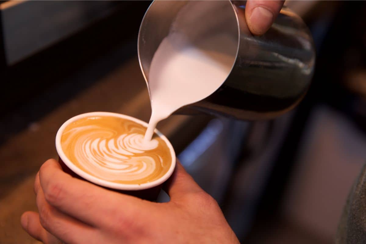 A dark close up photo of a person pouring homemade sugar-free creamer into their coffee cup with a rippled creamer effect on top of the coffee.