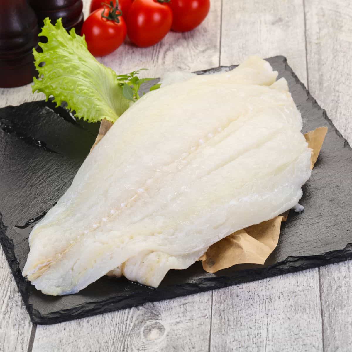 A raw piece of halibut fish filet on a black slab with a lettuce leaf and cherry tomatoes