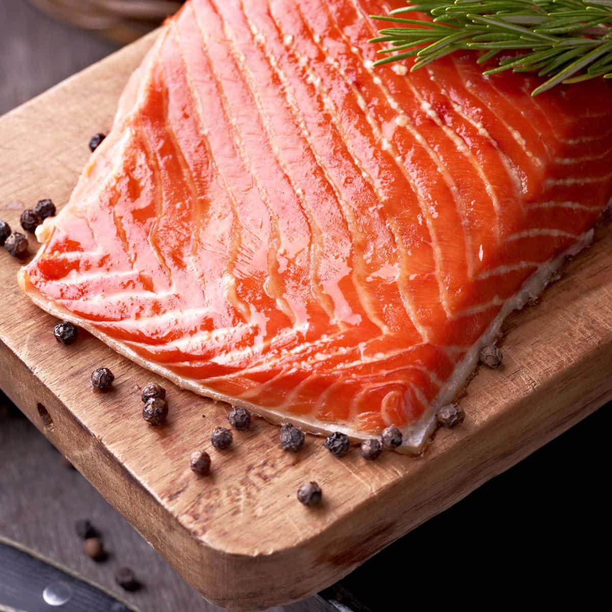 A raw filet of wild-caught salmon on a wooden slab surrounded by peppercorn decoratively