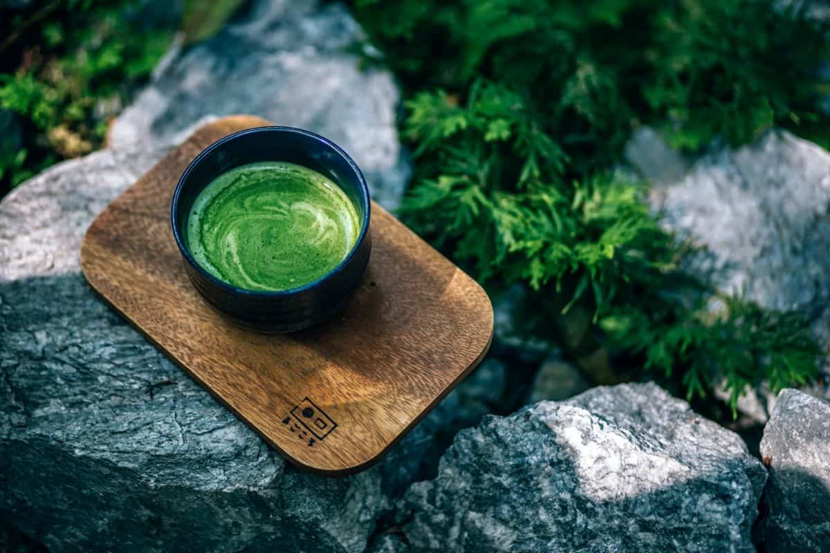 A cup of bright green matcha sits atop a wooden serving board on a mountain ledge with green trees around it.
