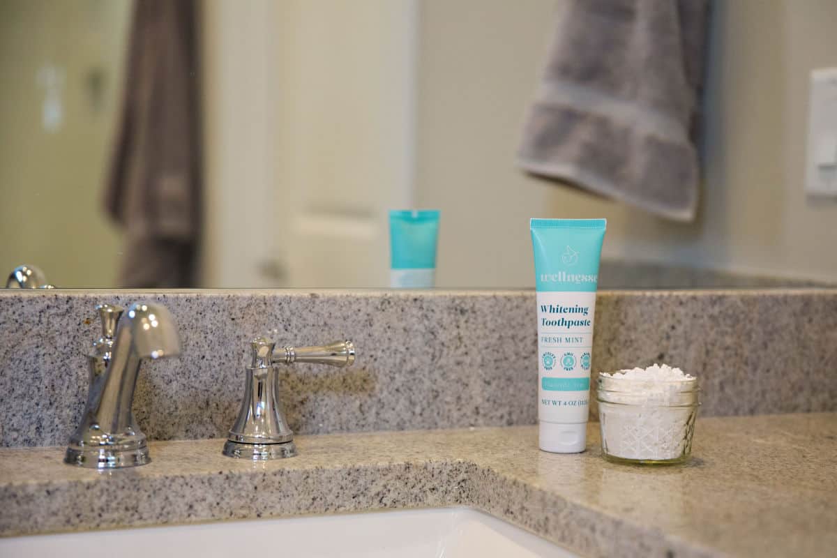 A close of of a bathroom sink and chrome faucet with a torquoise and white tube of toothpaste standing upright next to a jar. The tube says Wellnesse whitening toothpaste fresh mint fluoride-free.