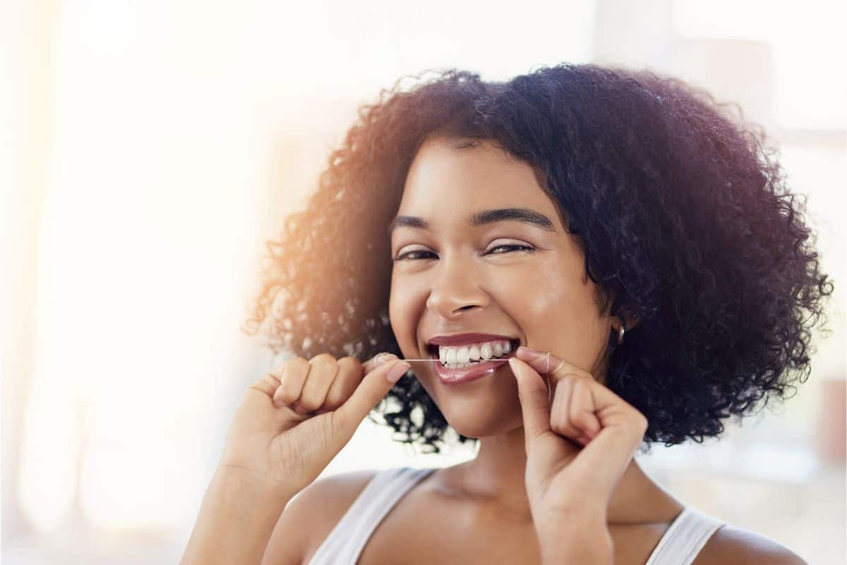 A woman with shoulder length naturally curly hair is smiling and holding a piece of dental floss next to her teeth