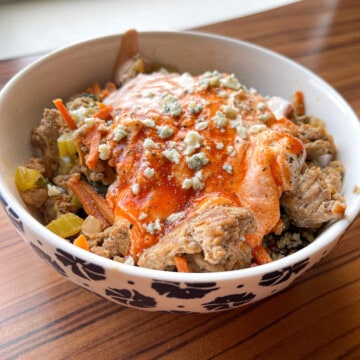 A white bowl with painted blue flowers on a wooden looking table. Inside the bowl is Buffalo Ground Turkey Quinoa Bowl with spicy yogurt sauce, blue cheese crumbles and black pepper seasoned on top.