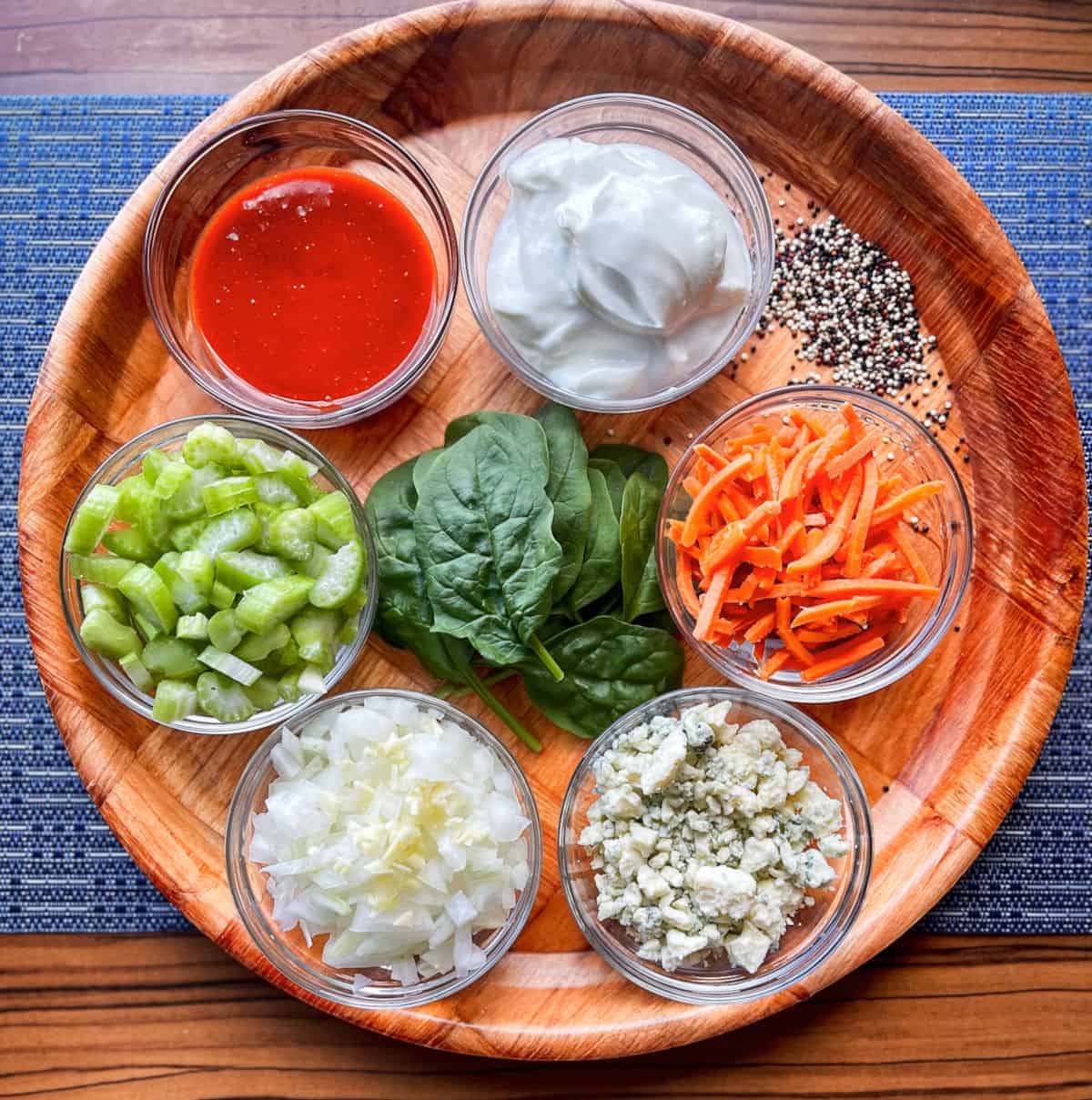 buffalo ground turkey quinoa bowl ingredients on a round wooden platter in clear glass ramekins and a pile of fresh baby spinach in the middle and dry tri colored quinoa off to the side. The ramekins have white ones minced, crumbled blue cheese, shredded carrots, minced celery, greek yogurt, and buffalo sauce in each of them. The platter is on a blue placemat.