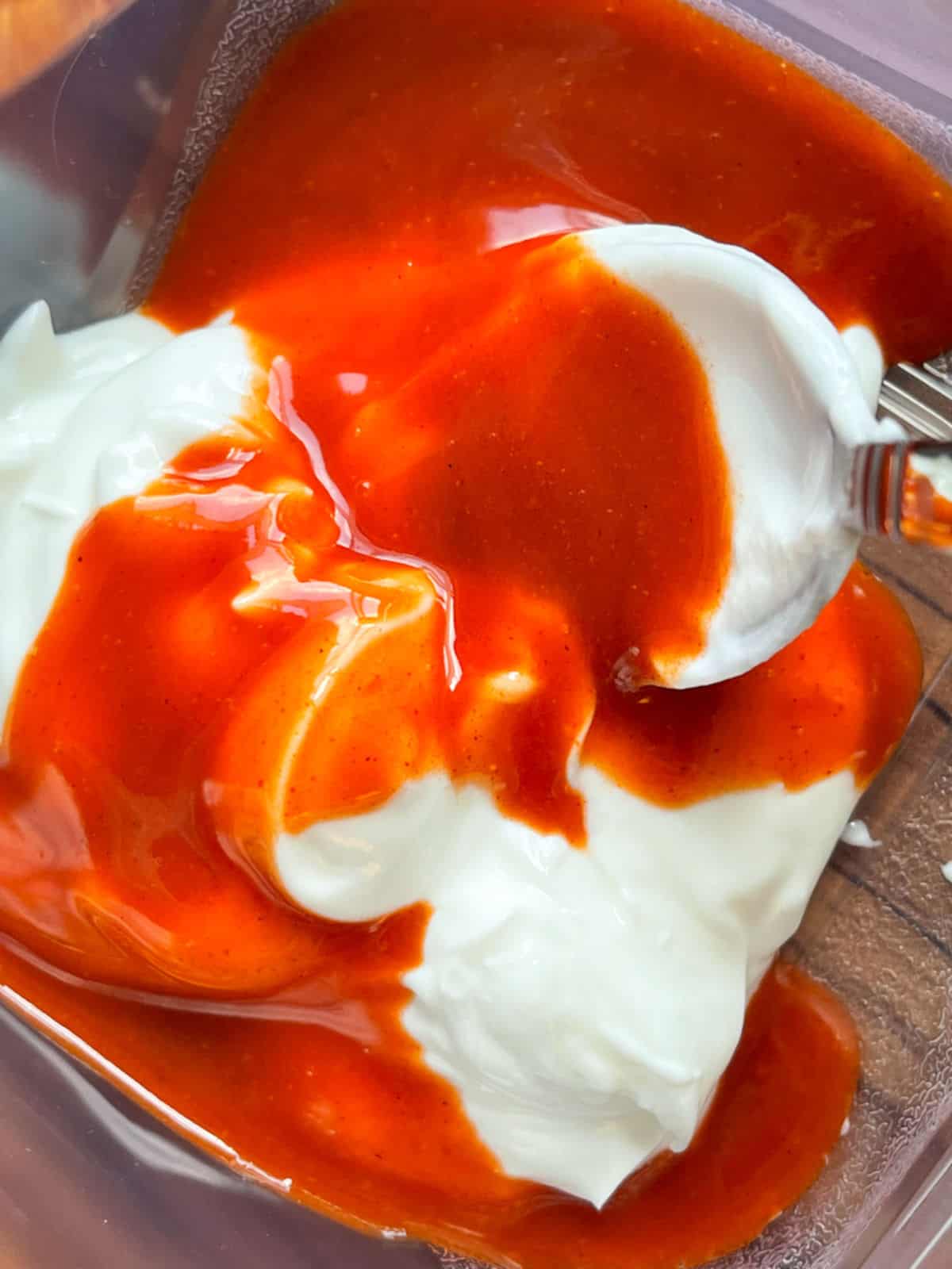 A clear bowl of greek yogurt with red buffalo sauce poured on top ready to mix the sauce.