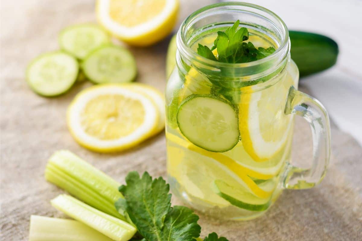 A clear glass mug with water, cucumber slices, lemon slices, and lime slices for detoxification support. Surrounding the cup is cut celery and more slices.