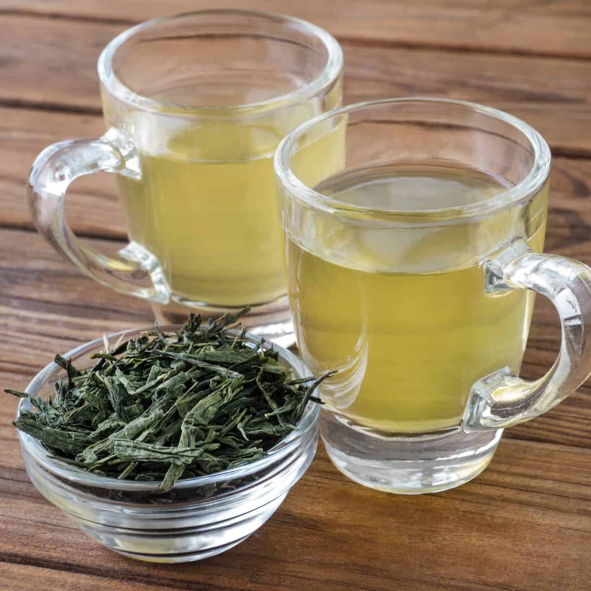 Two clear glass mugs with handles pointing in opposite directions. Each mug has a light greenish yellow colored tea in them with a bowl full of dried sencha leaves next to it.