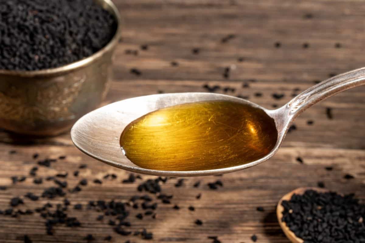 A close up of a spoon with golden black seed oil in it and black seeds scattered on a table in the background.