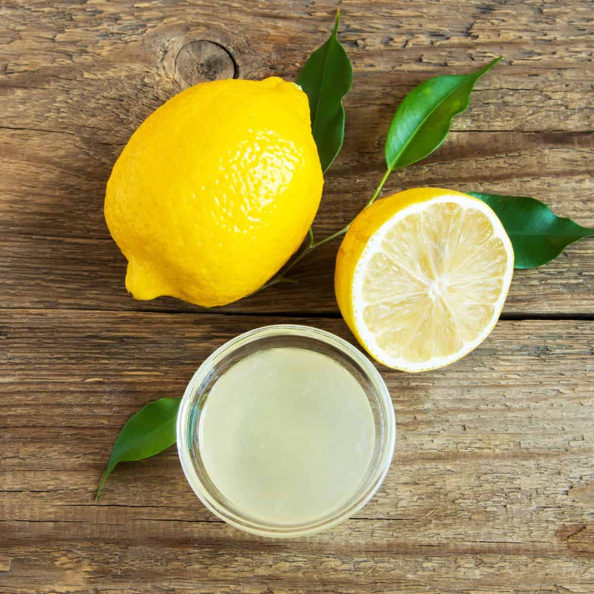 a wood background with one whole lemon with green leaves and one cut lemon next to a birds eye view of a glass of lemon juice