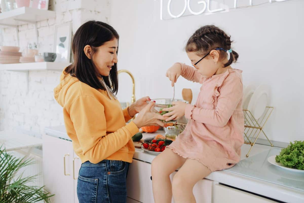 A mother has her child set on the counter top and she is teacher her to cook and eat healthy food in a bowl and on plates all around them are fresh fruit and vegetable options. The daughter has a spoon she is using to dig into the bowl the mother is handing her.