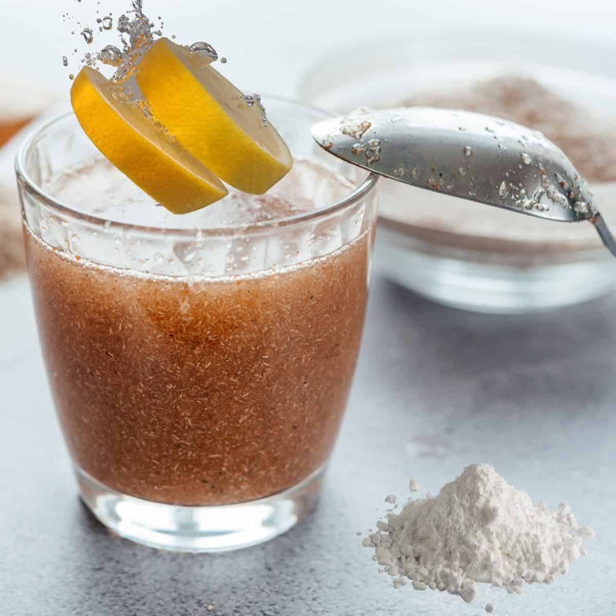 A cup of hydrated psyllium husk with a lemon slice splashing into it next to a pile of bentonite clay powder all in preparation for the mucoid plaque cleanse recipe