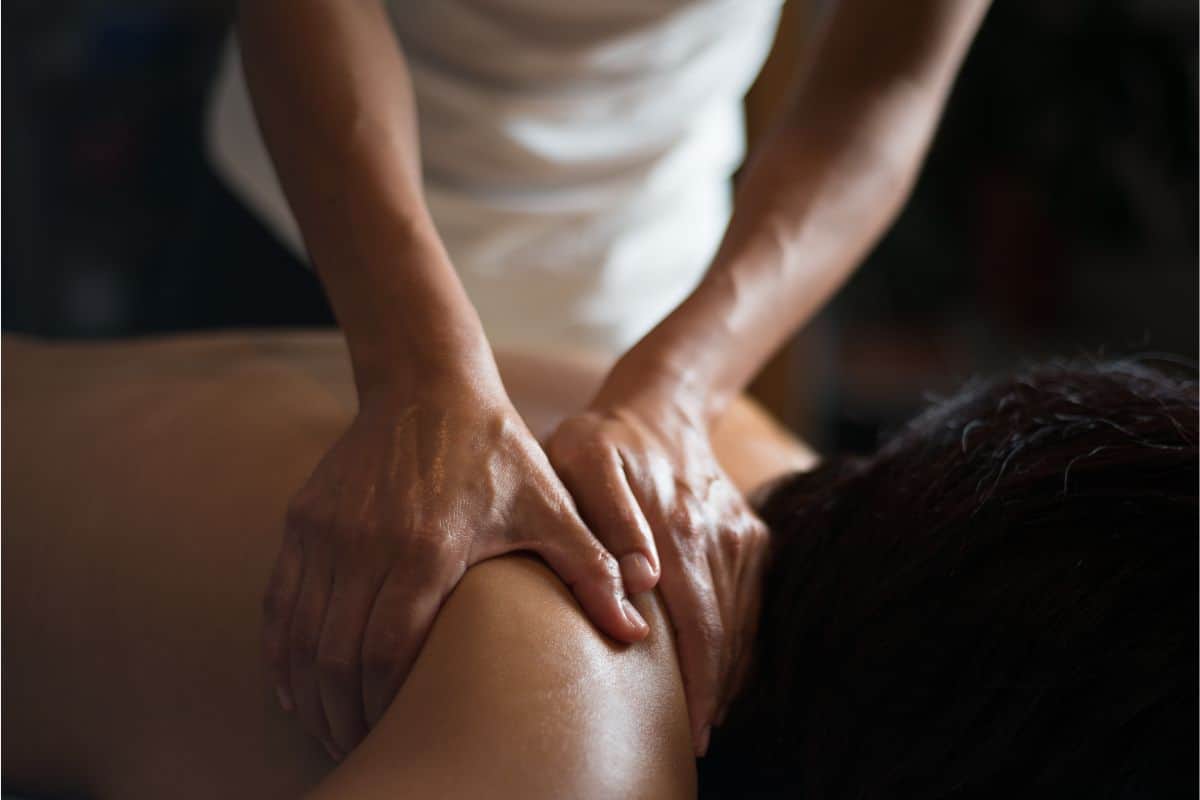 A photo of a massage therapist rubbing oil on a face down clients bare shoulder