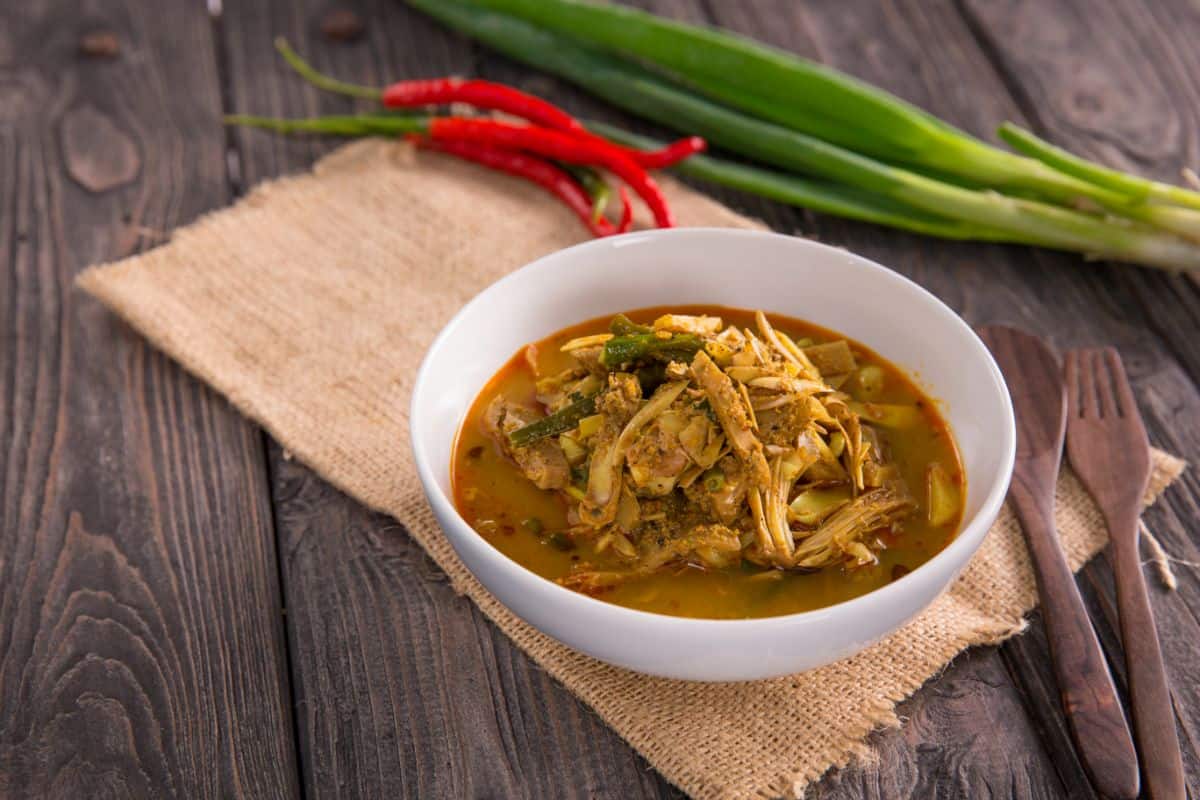 A photo of a white bowl with jackfruit curry soup in it mixed with vegetables and spices next to a burlap placemat and wooden utensils. In the background is a chili pepper cluster and a bundle of green onions. The meat of the jackfruit looks just like chicken or pork.